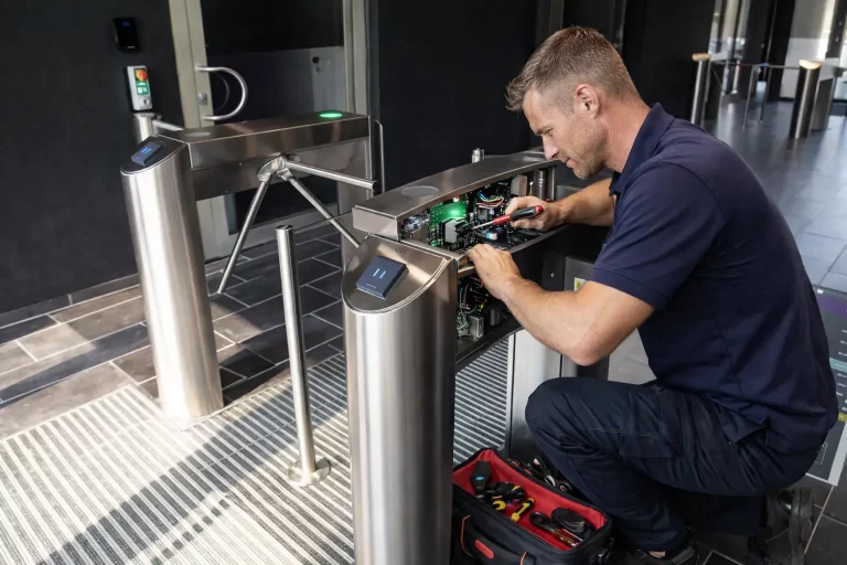 Ein Mann in einem marineblauen Hemd repariert die interne Elektronik eines Drehkreuzes in einer modernen Gebäudehalle. Er hockt neben einer offenen Schalttafel und benutzt Werkzeuge aus einer rot-schwarzen Werkzeugtasche auf dem Boden.