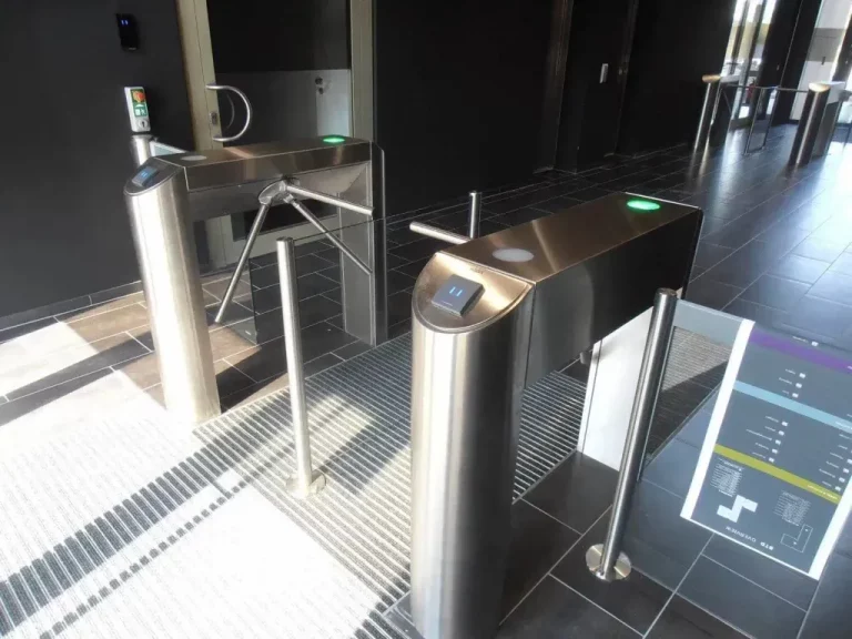 Zwei Drehkreuze aus Edelstahl mit Kartenlesegeräten befinden sich an einem Eingang in einem modernen Gebäude mit schwarz gefliestem Boden und minimalistischem Dekor.